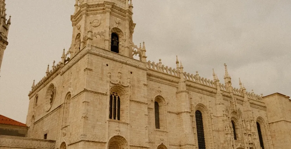 Ornate beige limestone facade of Jerónimos Monastery with a bell tower and arched windows, people walking in front.