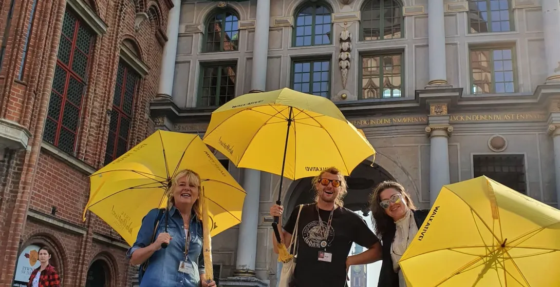Three smiling Walkative guides hold yellow umbrellas on a cobblestone street in front of Gdańsk's historic Green Gate buil...