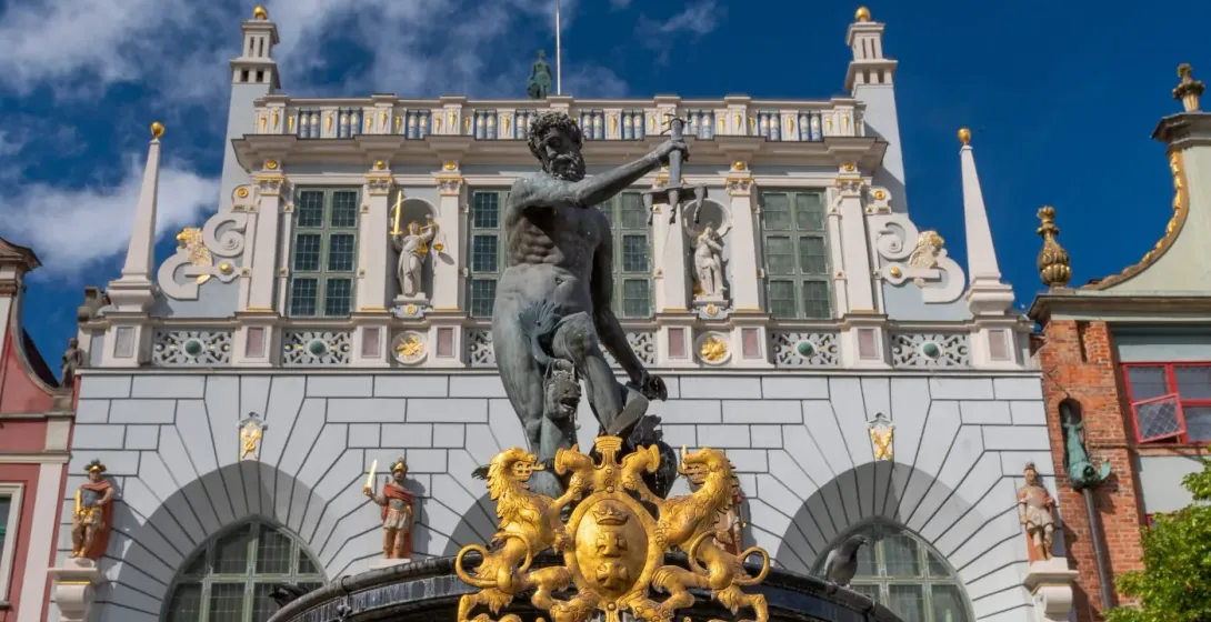 Bronze statue of Neptune with a trident on a fountain, in front of a grand, ornate building with a light grey facade.