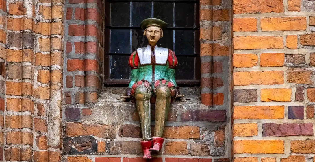 Colorful wooden figure of a man in a green beret and medieval-style clothes, sitting on a brick window sill of an old buil...
