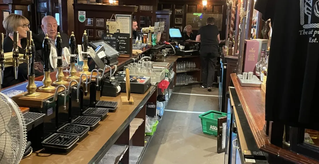 Interior of a traditional Glasgow pub with a long wooden bar, many beer taps, and banknotes from various countries strung ...