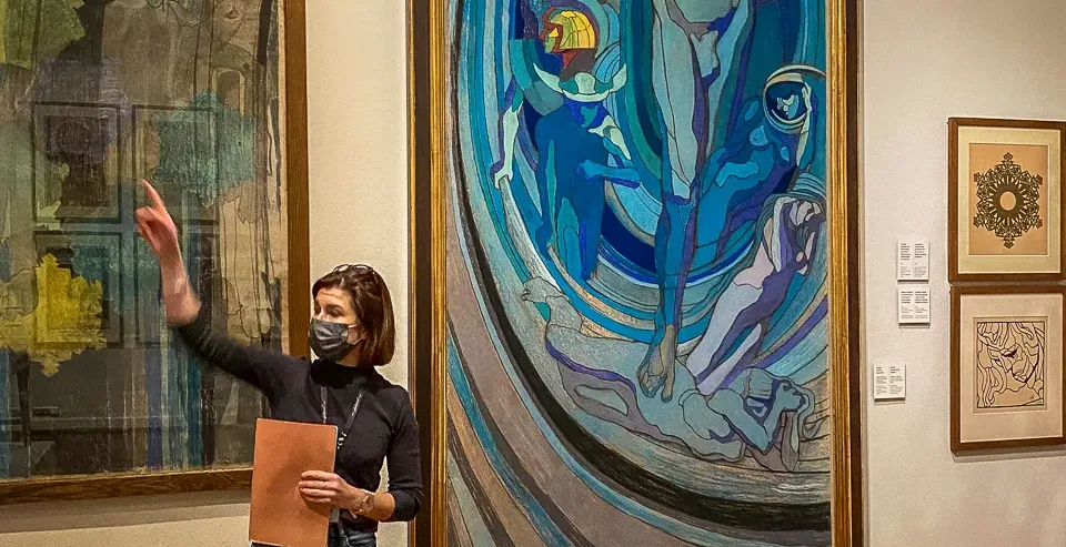 A tour guide wearing a face mask stands in an art gallery, pointing towards a large colourful painting on the wall.