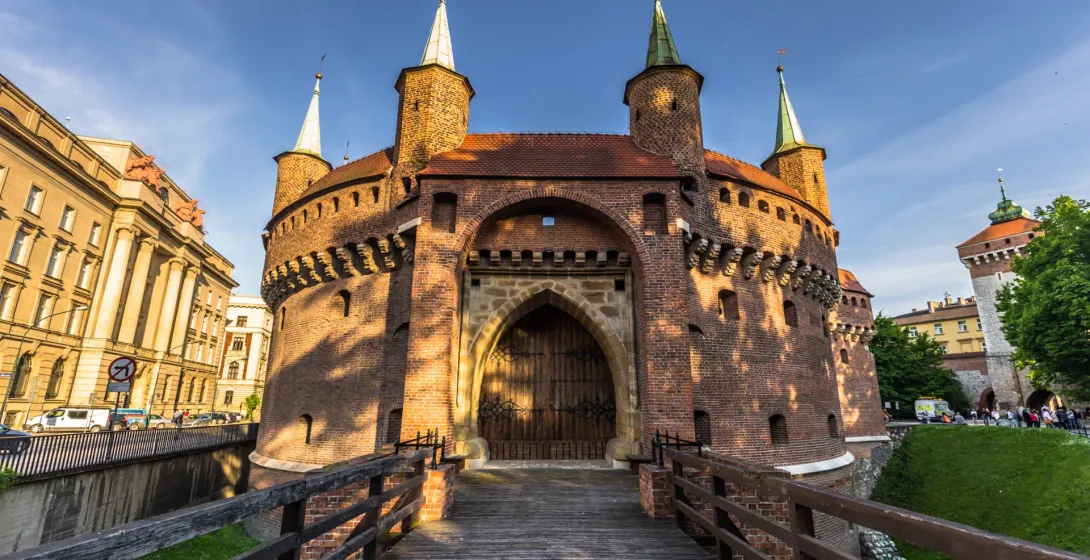 The Krakow Barbican, a circular red-brick fortress, seen from its wooden entrance bridge on a sunny day.