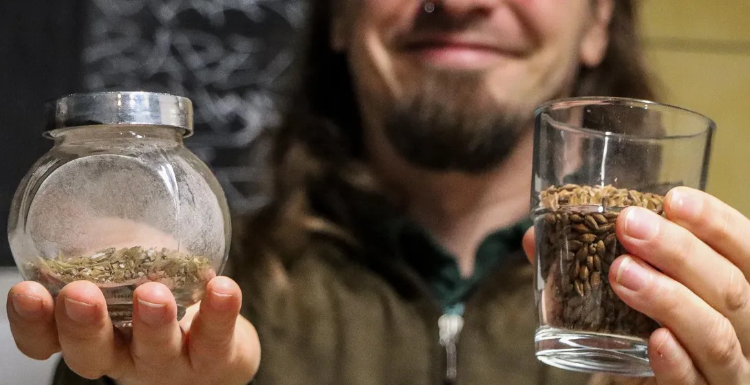A man with long hair holds up a glass of malted barley and a small jar of dried hops toward the camera.