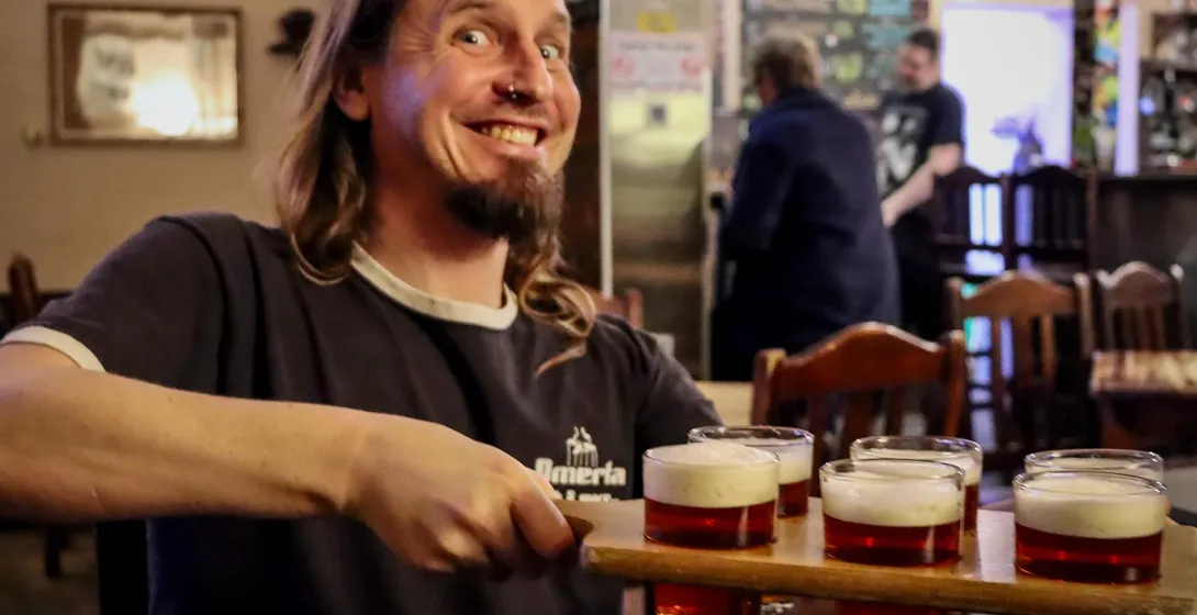 A smiling man with long hair and a nose ring holds a wooden flight with six small glasses of beer in a pub.