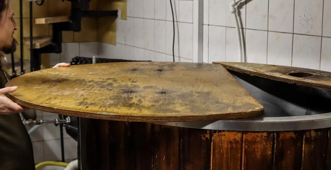 A brewer opens the wooden lid of a large brewing vat in a small brewery.