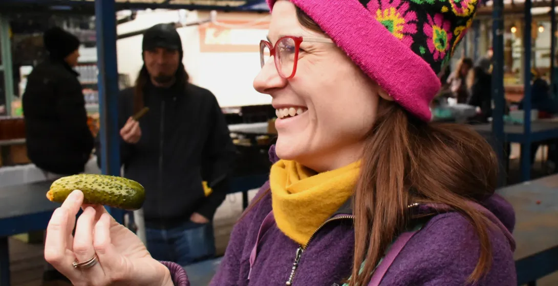 Smiling woman wearing a colorful beanie holding a traditional pickled cucumber at an outdoor food market in Krakow.