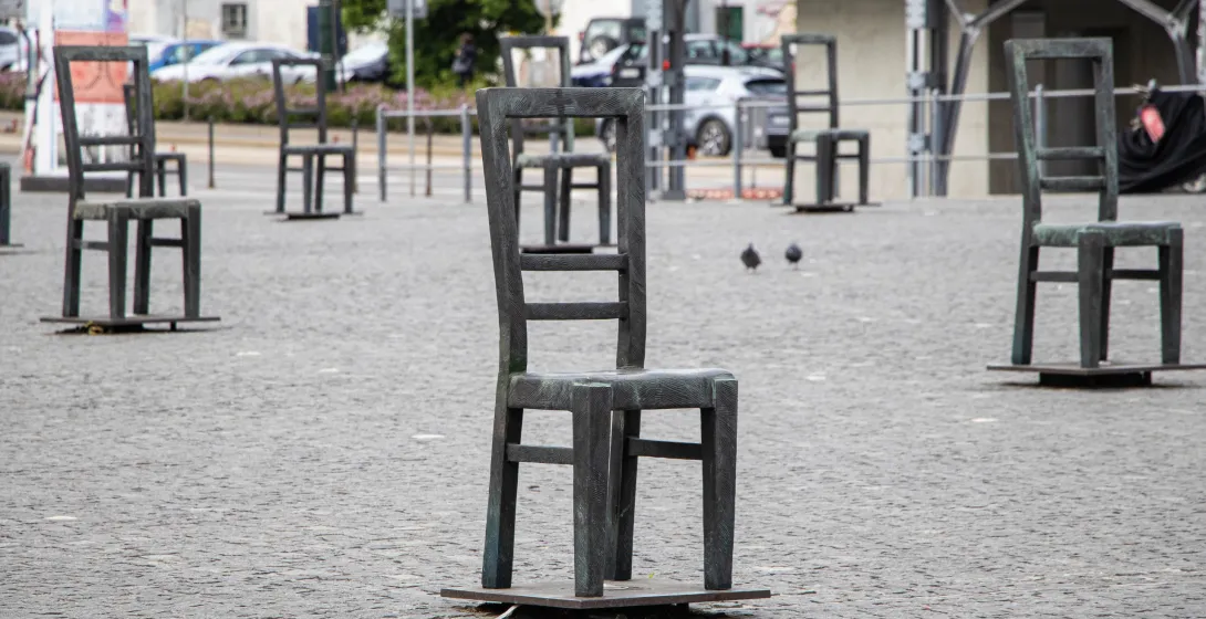 Metal chair sculptures fill a cobblestone square, with two pigeons visible and buildings in the distance.