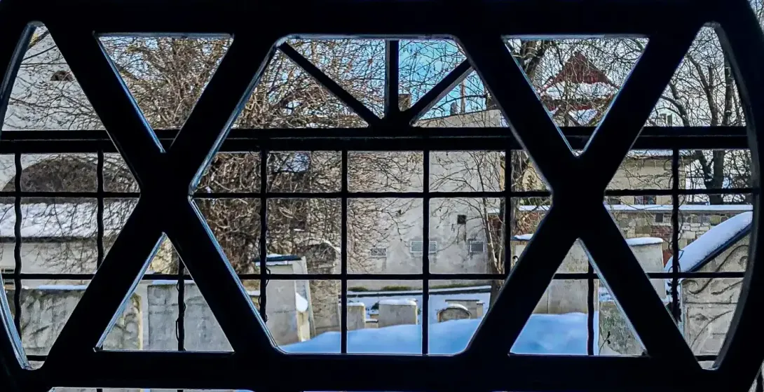 View through a window with a Star of David grille. Outside, a snow-covered cemetery with gravestones and bare trees.
