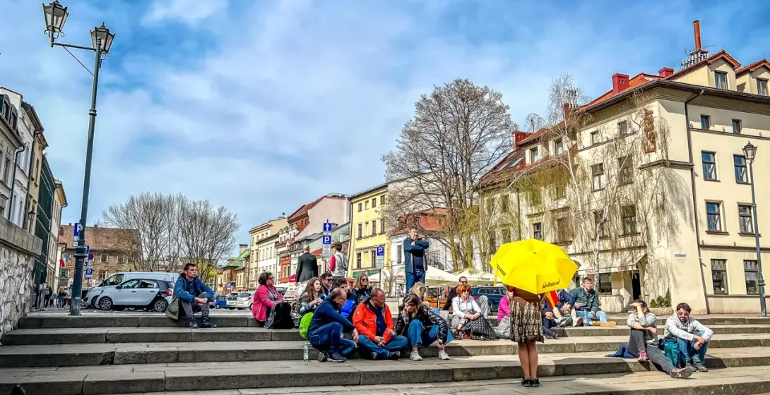 People sit on wide stone steps in a city square, historic buildings and a lamppost under a partly cloudy sky. One person h...