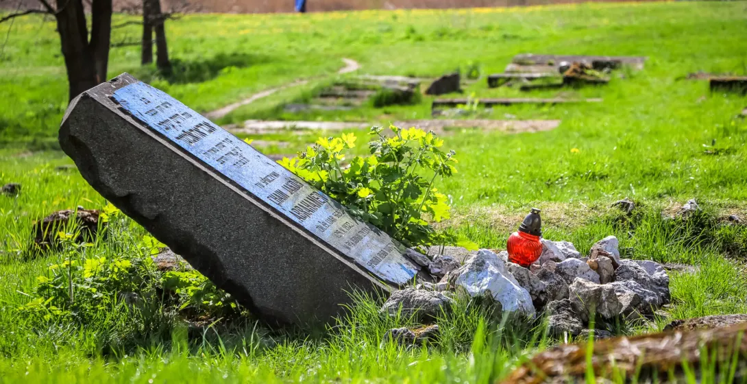 An inclined stone memorial with inscriptions stands in a green field, surrounded by small rocks and a red glass candle.