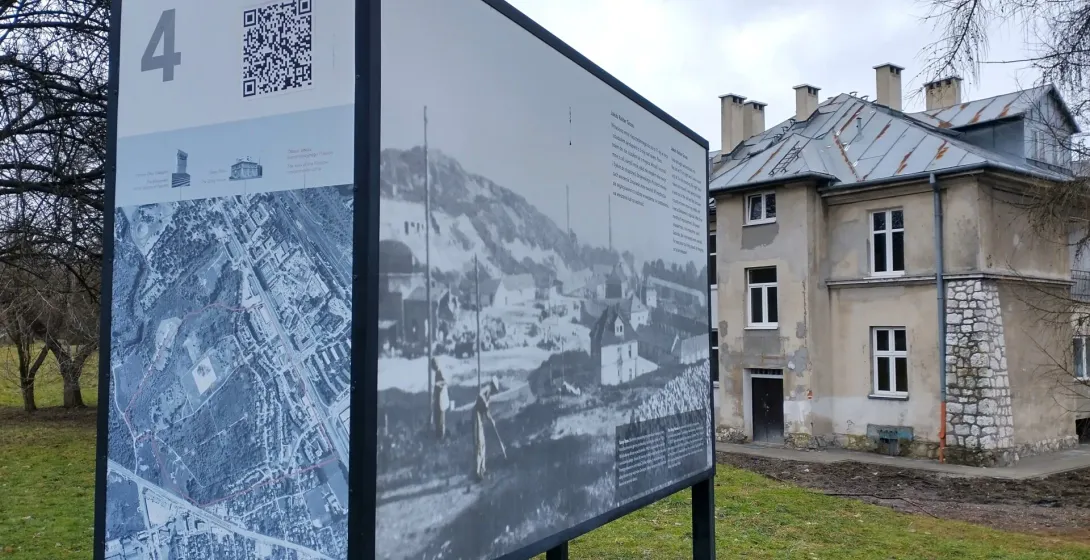 An information board with a map and historical photos stands on a lawn in front of the 'Grey House' at the Plaszow camp site.