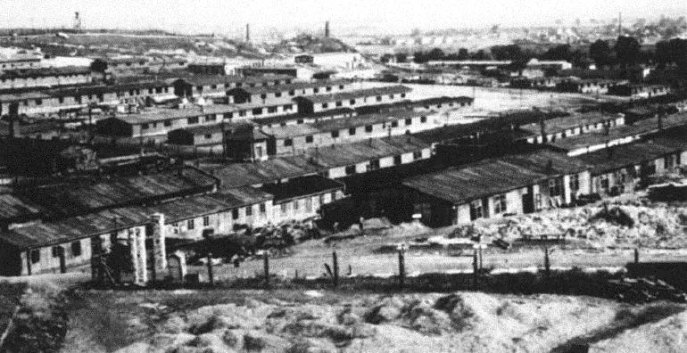 Black and white aerial view of Plaszow Concentration Camp with numerous barracks, fences, and distant industrial structures.
