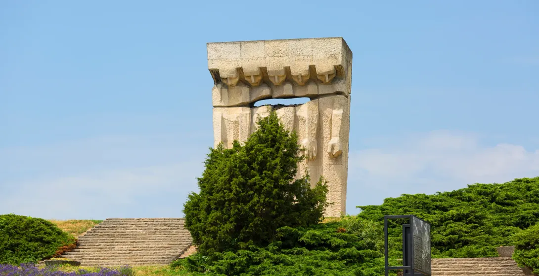 A large, abstract stone memorial monument stands against a clear blue sky, surrounded by green shrubs, stone stairs, and p...