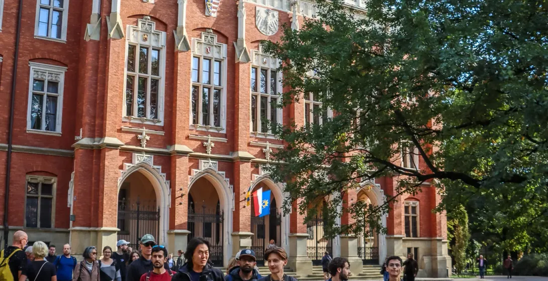 A tour group walks past the red brick neo-Gothic facade of the Collegium Novum in Krakow on a sunny day.