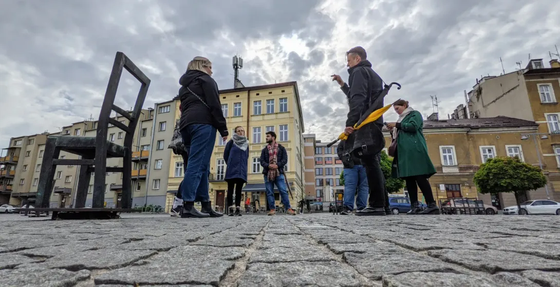 A tour guide speaks to a small group on a cobblestone square with large metal chair sculptures and buildings under a cloud...