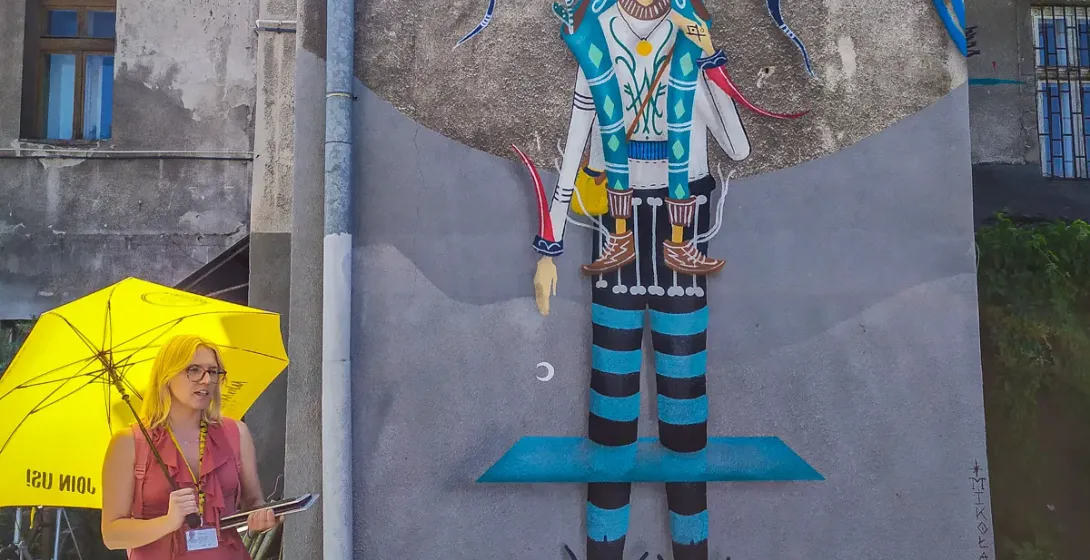 A tour guide with a yellow umbrella stands in front of a large, colorful mural on a building in Krakow.
