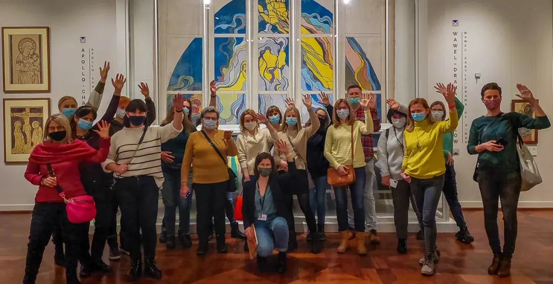 A group of tourists wearing face masks waves inside a museum in front of a large, modern artwork.