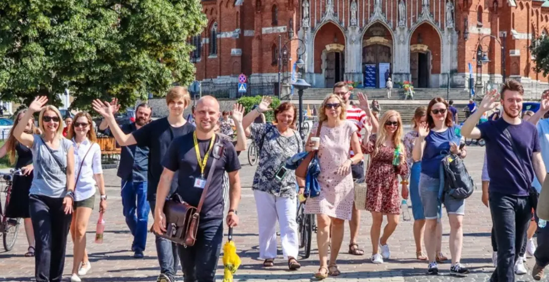 A tour guide with a yellow umbrella leads a group waving at the camera in front of a large brick church in Krakow.