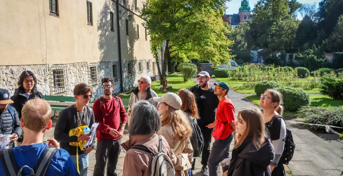 A group of tourists listening to a guide on Wawel Hill in Krakow, with the cathedral towers visible in the background.