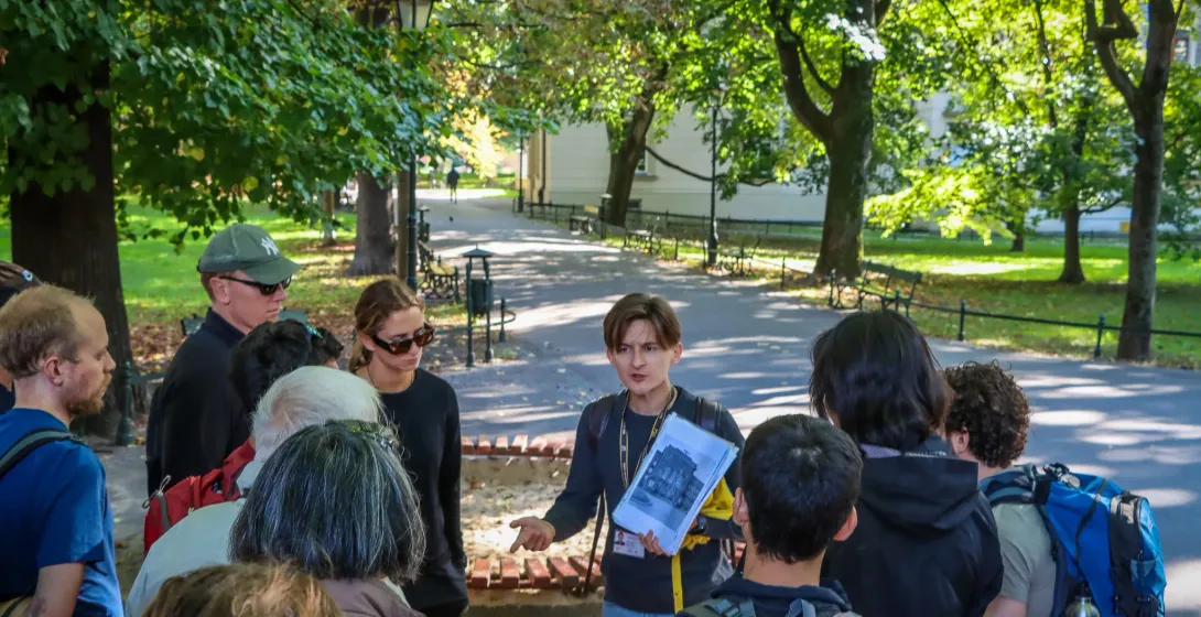 A tour guide shows historical photos to a group of people on a path in a sunlit park in Krakow.