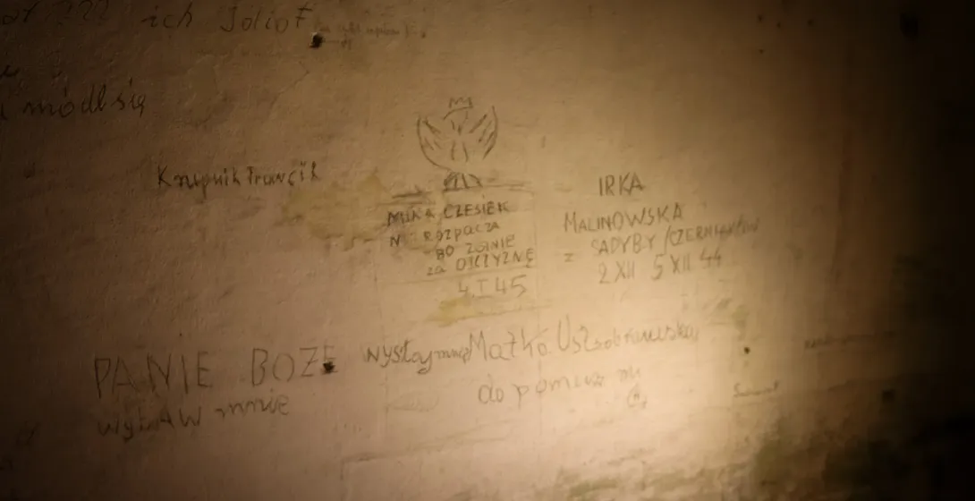 Dimly lit wall with pencil inscriptions in Polish from the WWII era, including a hand-drawn Polish eagle with a crown.