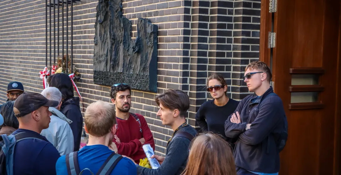 A tour guide explains a point to a group of travellers in front of a memorial plaque on a brick building in Krakow.