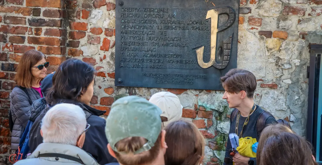A tour guide explains a historical plaque with the Polish Fighting Symbol to a group of tourists against a brick wall.