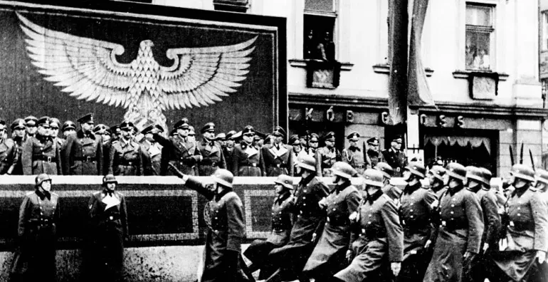 Black and white photo of a Nazi military parade on a street in Krakow. Soldiers march past a stand with officers and a lar...