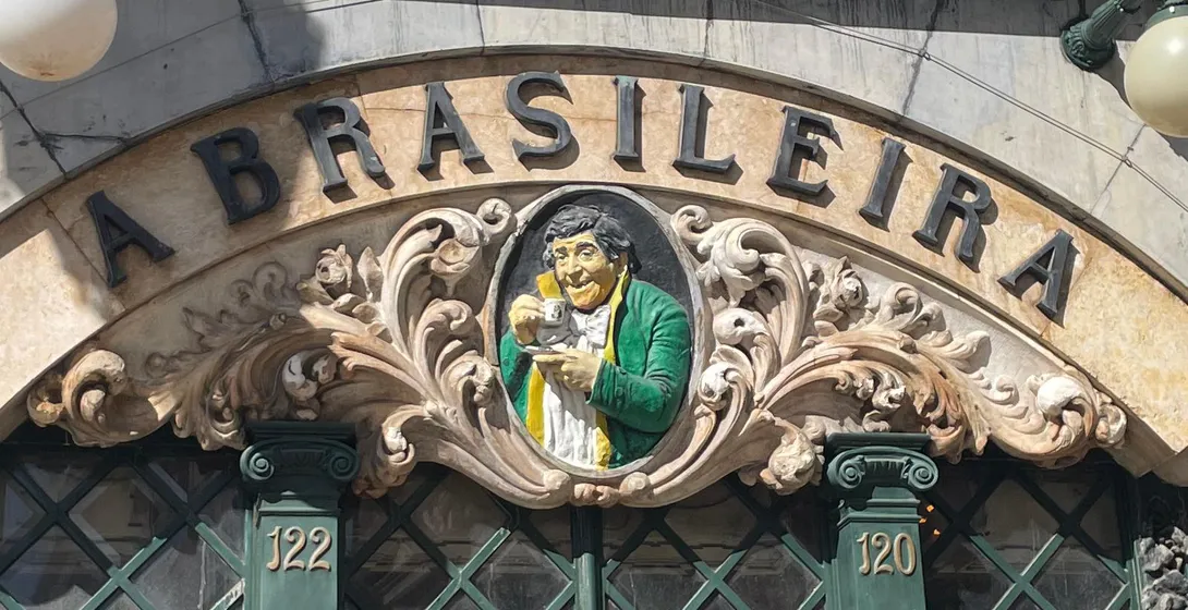 Facade of Café A Brasileira in Lisbon, showing its arched sign, a central relief of a man with a cup, and green-framed win...