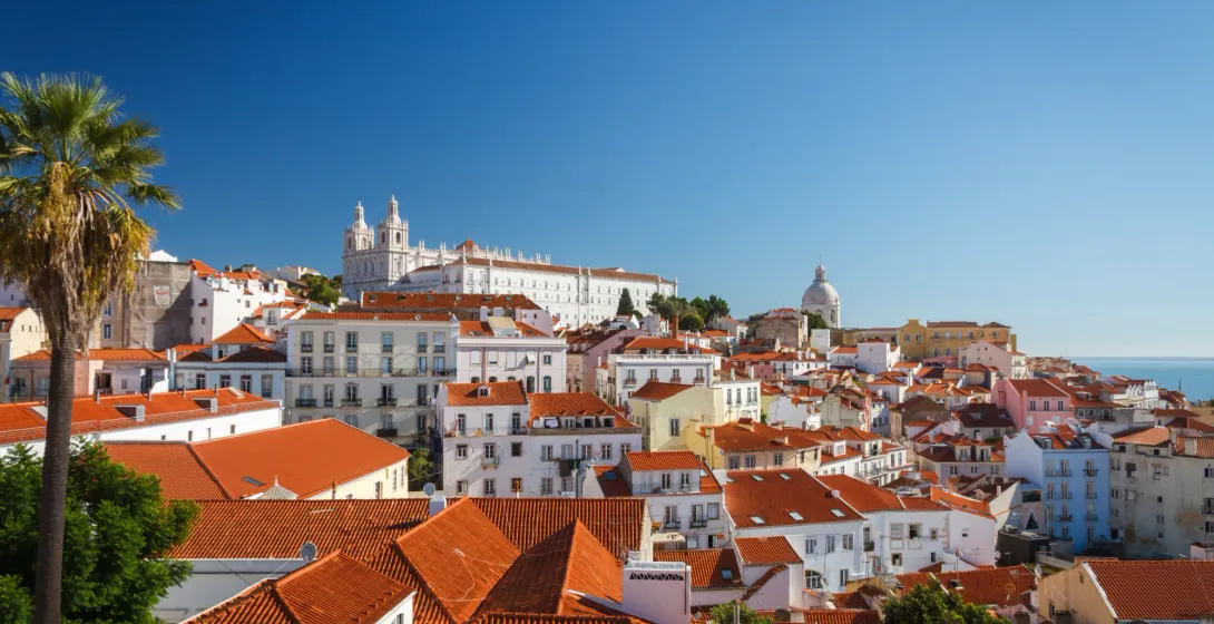 Red tile roofs of Lisbon's Alfama district with the white São Vicente de Fora Monastery under a clear blue sky.