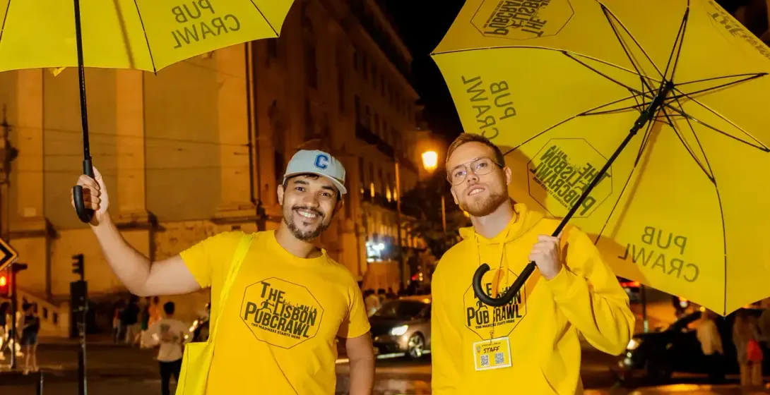Two men in yellow 'The Lisbon Pubcrawl' apparel holding yellow umbrellas, standing on a wet street at night in Lisbon.