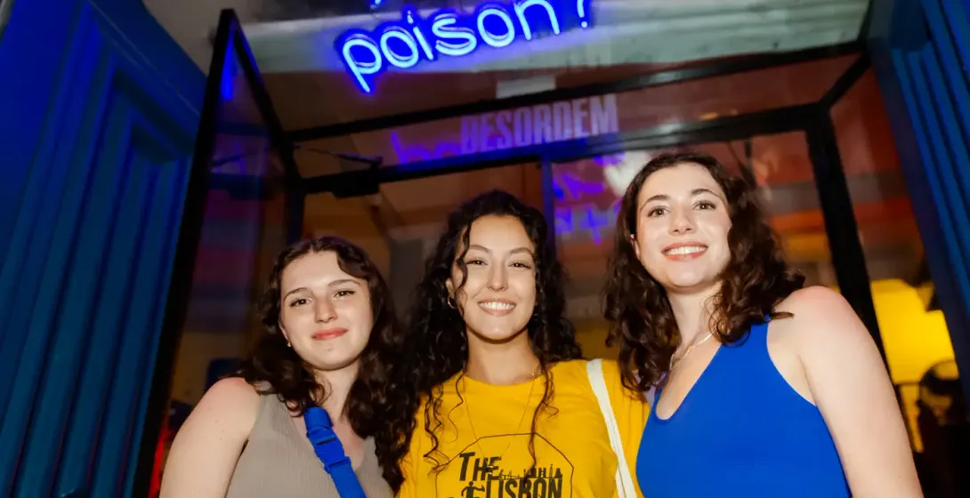 Three young women smiling in front of a blue neon sign that reads 'What's your poison?' above a blue door frame.