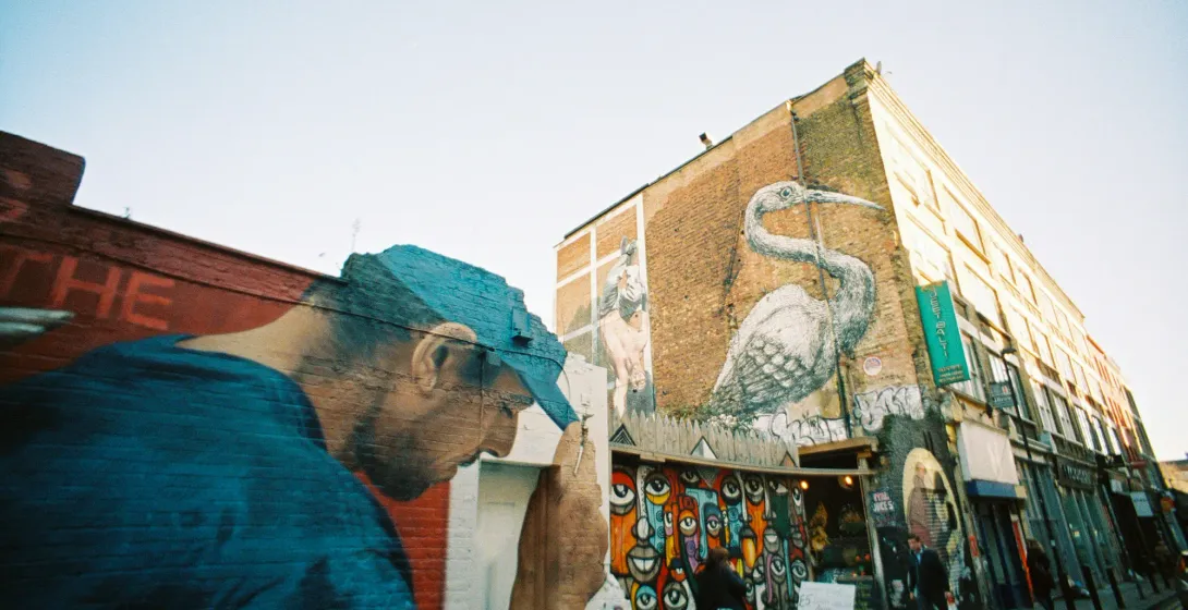 A London street with large-scale murals on brick buildings, including a large white heron and other colourful artwork.