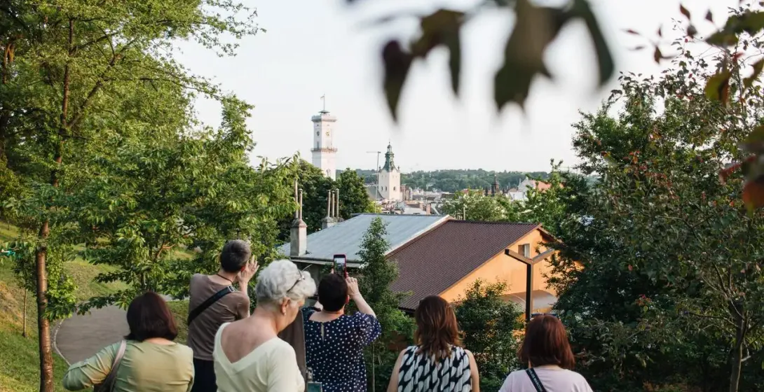 Group of people on a hill overlooking Lviv cityscape with City Hall tower and a church dome visible through trees.