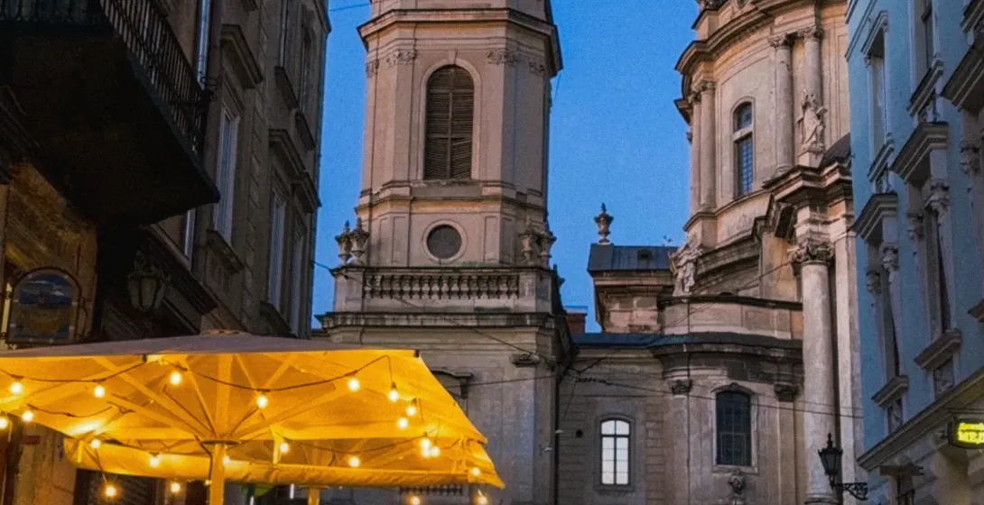 A busy street cafe in Lviv at dusk, with the Dominican Cathedral in the background under a deep blue sky.