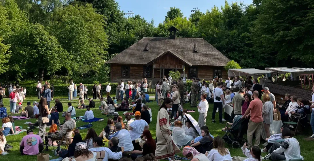 A large crowd of people gathered on a grassy lawn in front of a traditional wooden house on a sunny day.