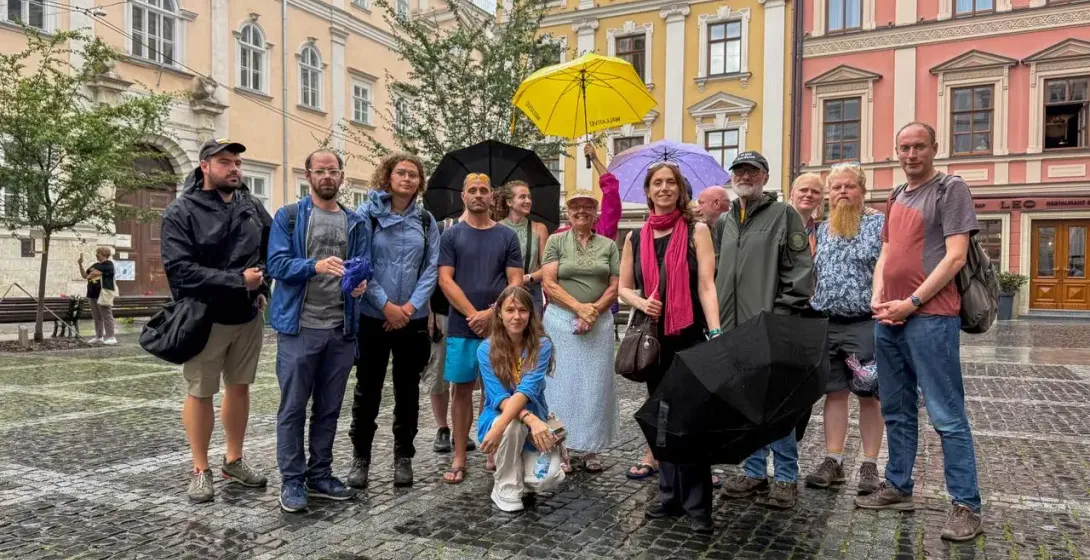 A group of about 15 adults stands on a wet cobblestone square in Lviv. Several hold umbrellas, with colorful old buildings...