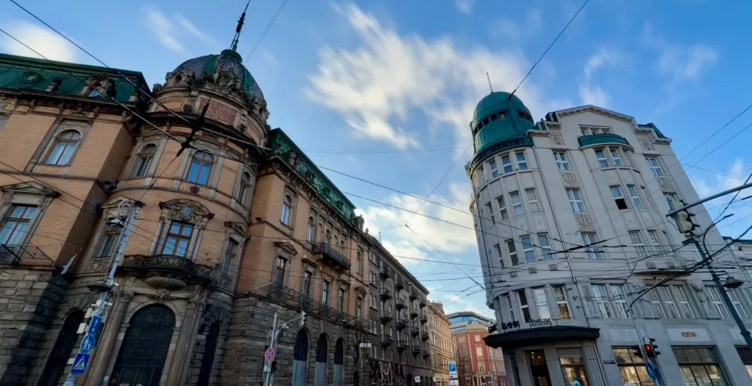 Wide-angle view of two historic buildings on a wet cobblestone street in Lviv under a blue sky with streaked clouds.