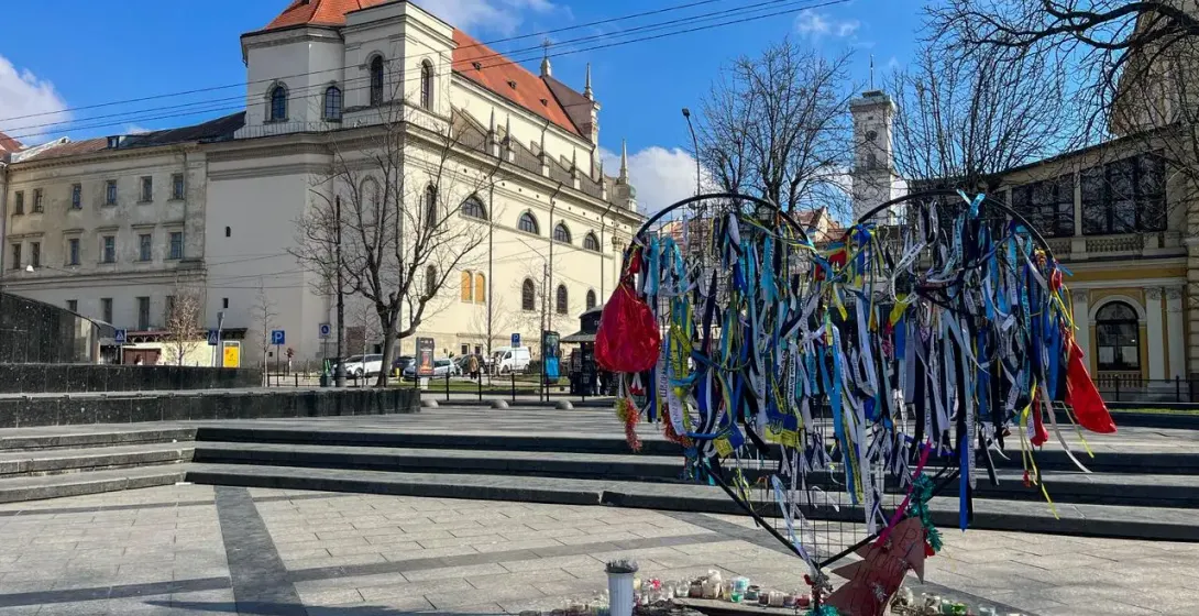 Heart-shaped memorial frame decorated with blue and yellow ribbons and many candles in a Lviv square, with a historic buil...