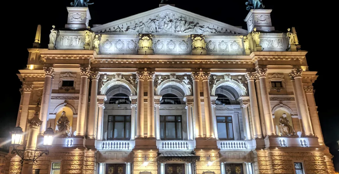 The brightly illuminated Lviv Opera House at night, with detailed sculptures and people walking in front.