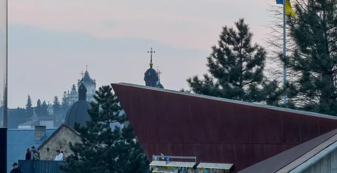Lviv skyline with church domes, a modern building, people on a path, and a Ukrainian flag at dusk.