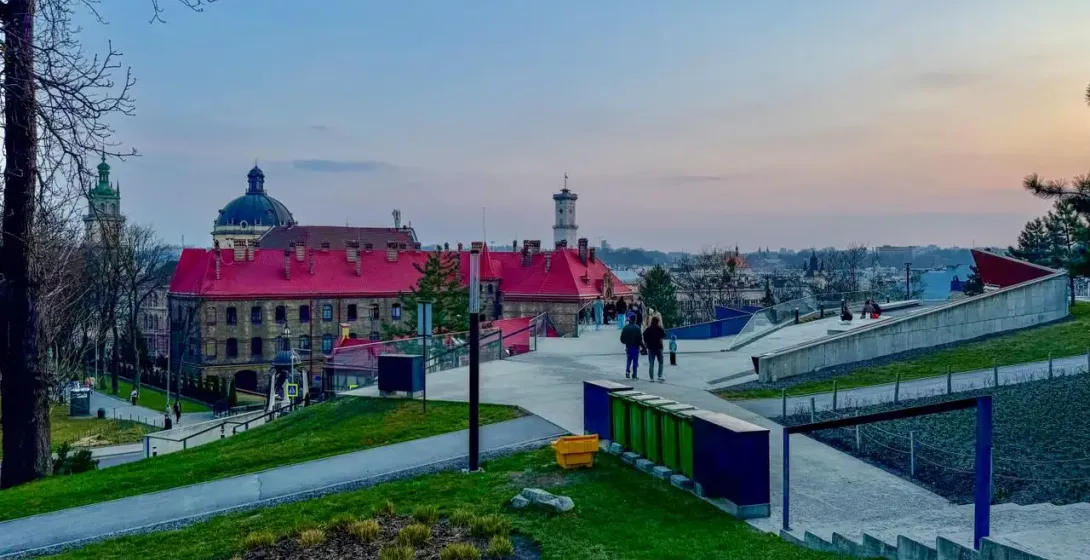 View from High Castle Hill in Lviv at sunset, showing red-roofed buildings, a dome, a tower, and modern park paths with pe...