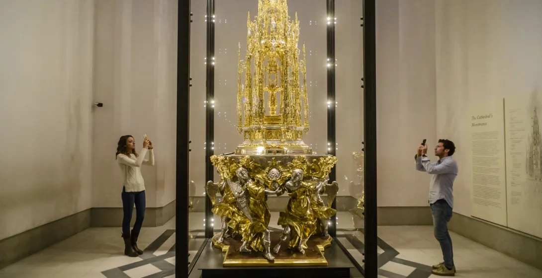 An ornate, gilded monstrance in a glass display case, being photographed by two visitors on their phones.