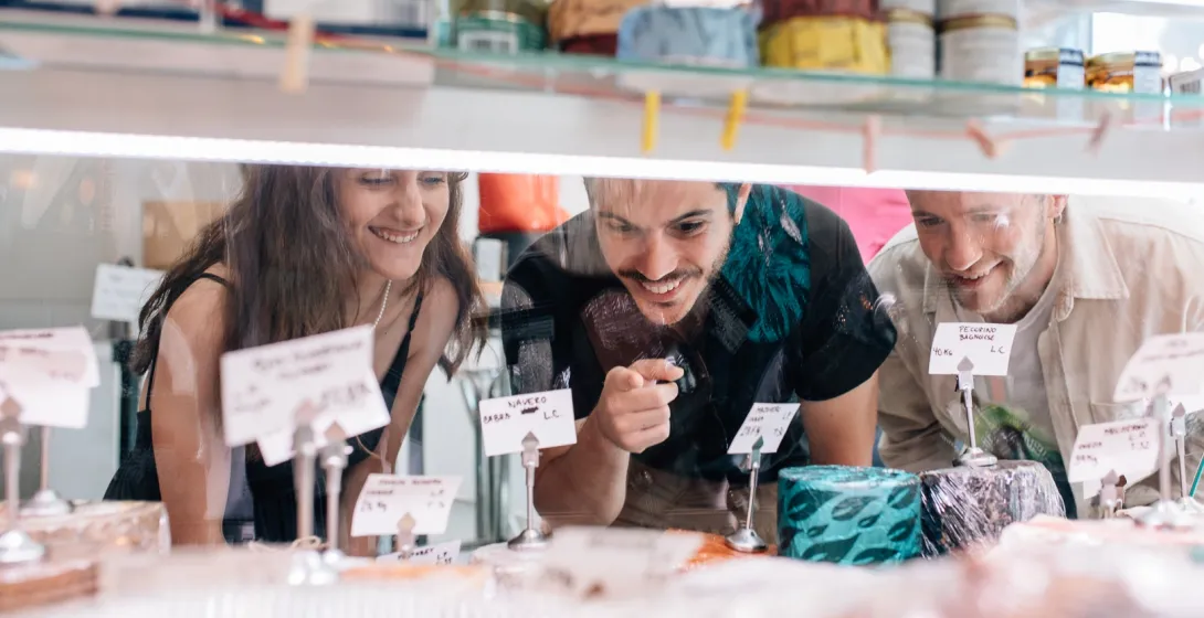 Three young people smile as they look through a glass counter at a selection of cheeses in a shop.