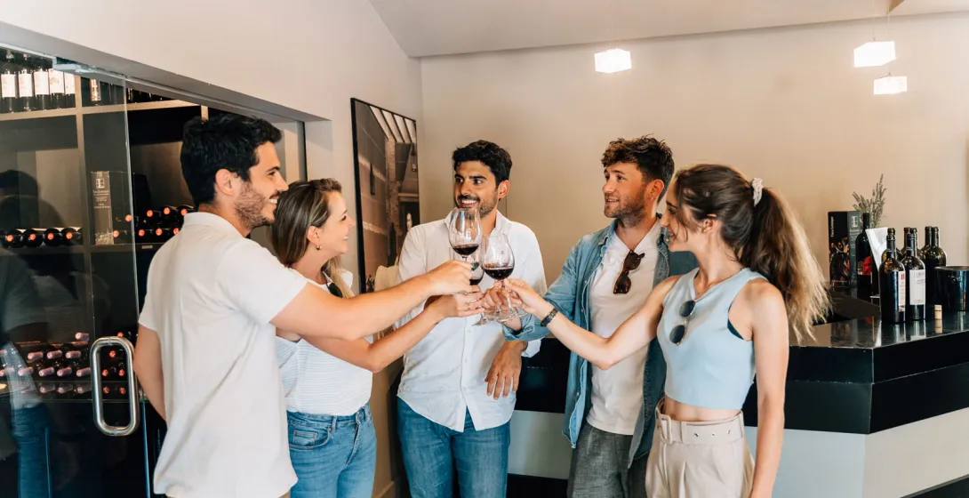 A group of five young adults toasting with glasses of red wine inside a modern wine shop.