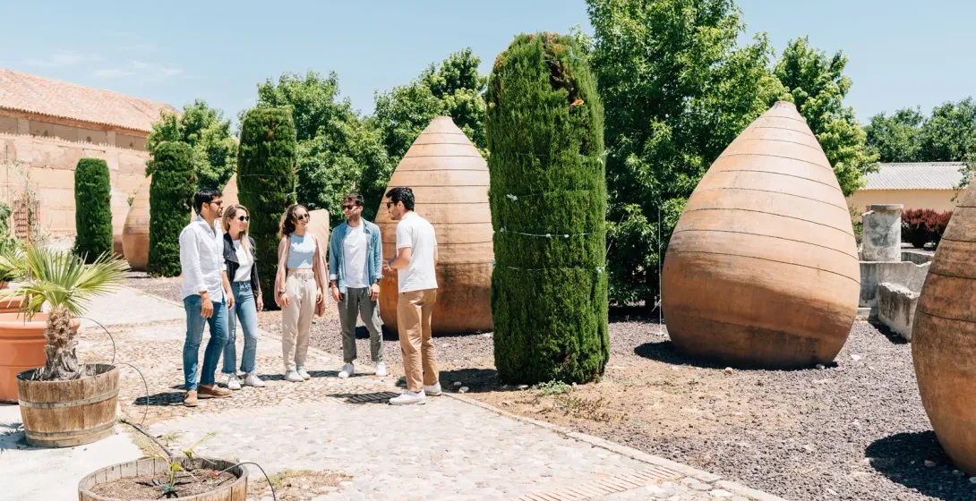 A tour guide speaks to a small group of people in a sunny courtyard with large, ancient-style clay jars.