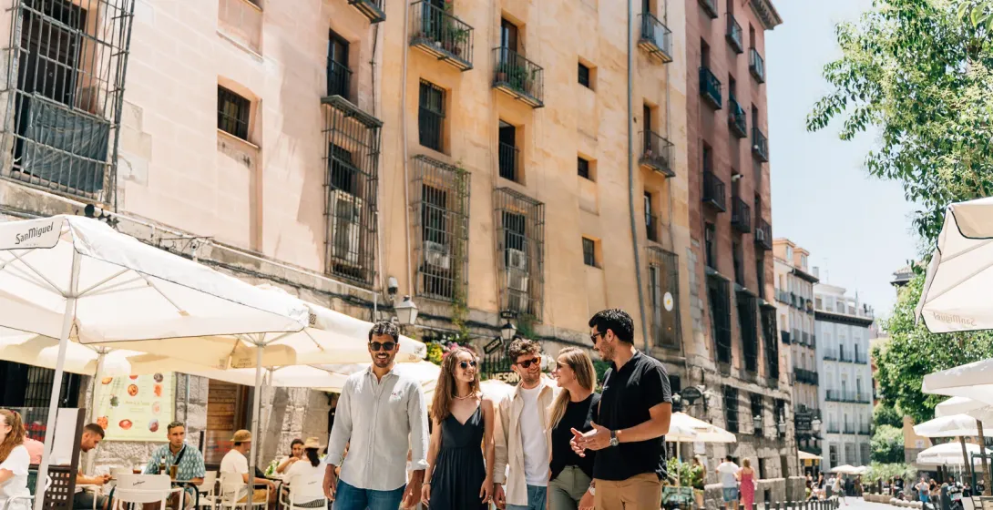 A group of five young people walk down a sunny Madrid street lined with cafe tables and historic buildings.