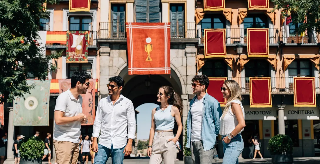 A group of five young adults in a sunny plaza, in front of a building decorated with red and gold banners.