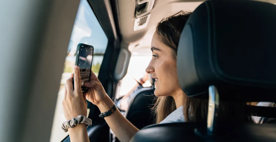 A young woman sits in the back of a car and takes a photo of the view with her smartphone.